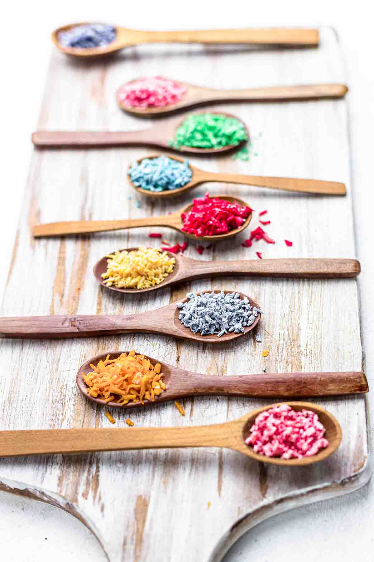 Colorful sprinkles in wooden spoons on a wooden cutting board.