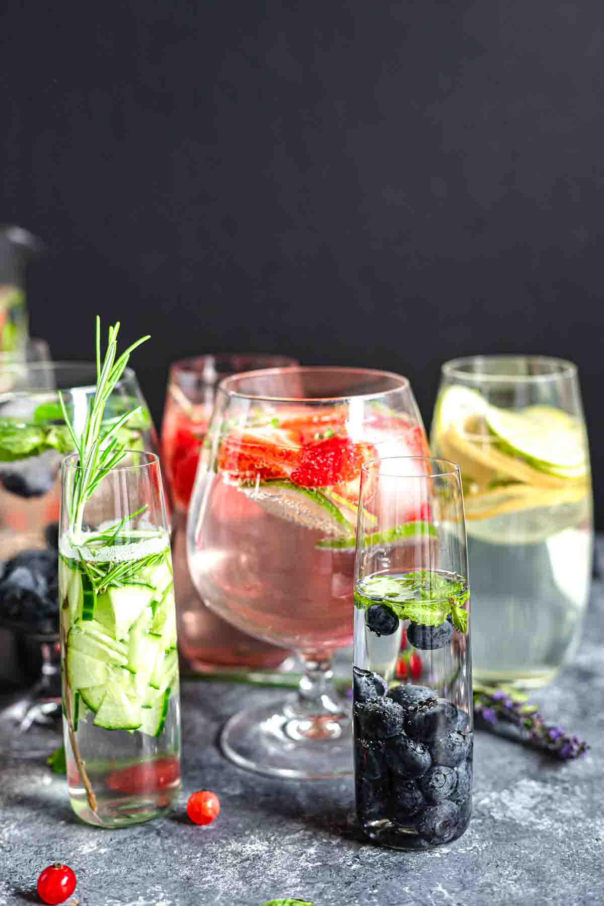 Assorted glasses of infused water, each containing different fruits, herbs, and berries, arranged on a dark background.