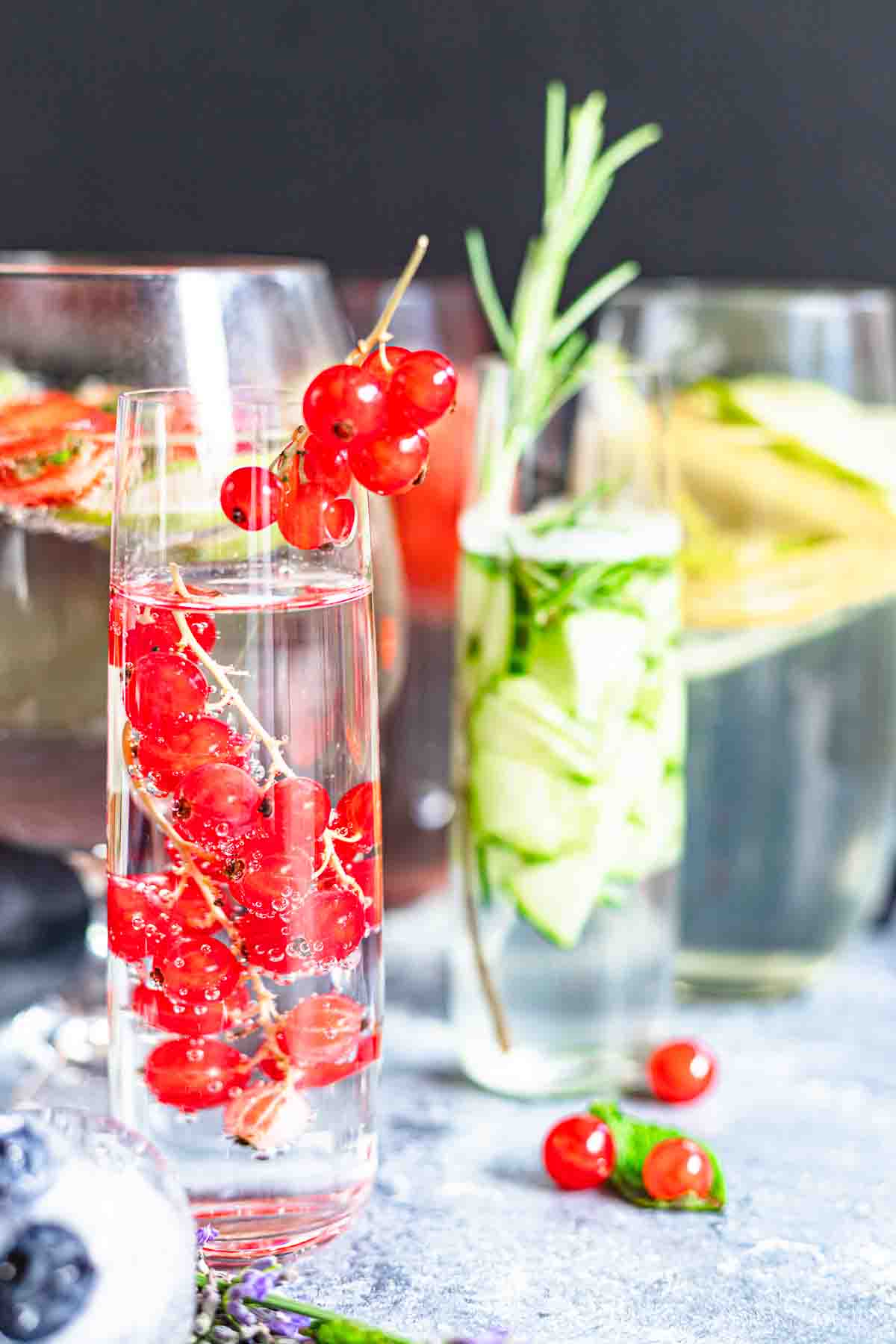 Three different fruit infused waters in tall glasses, garnished with fruits like red currants, strawberries, and cucumbers, arranged on a textured table.