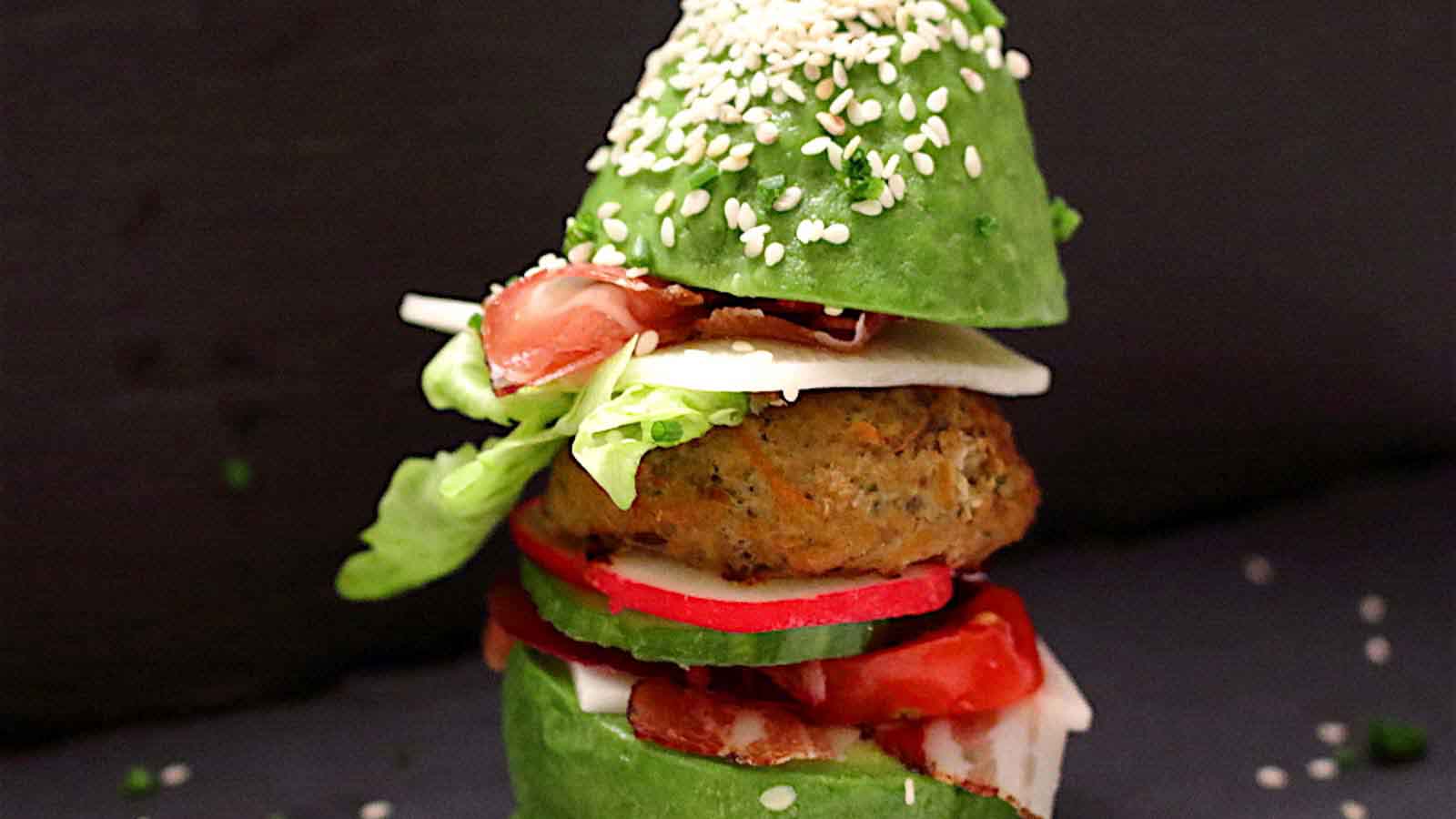 An avocado burger with a patty, lettuce, radishes, tomatoes, and cheese, topped with a sesame-seeded avocado half.