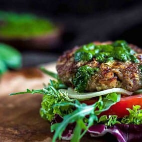 A close-up of a lettuce-wrapped burger patty topped with green sauce and layered with tomato, onion, and additional lettuce on a wooden serving board.