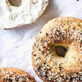 Several everything bagels, one sliced and spread with cream cheese, arranged on a white parchment paper.