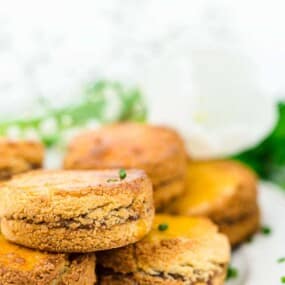 A close-up of a stack of fresh-baked scones, garnished with green herbs, placed on a white surface with a blurred background of a flower and a bowl of white dip on the side.