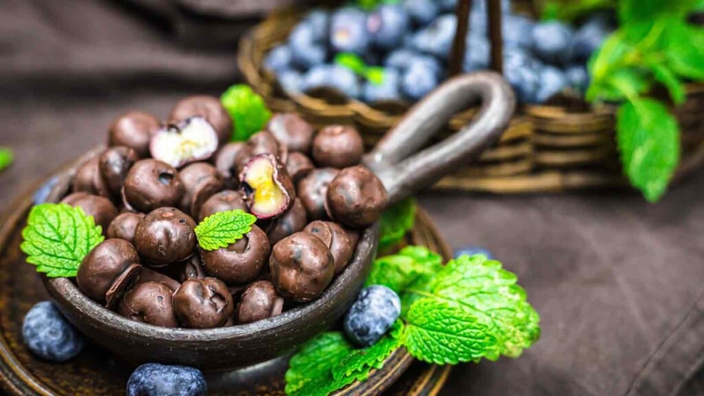 A small bowl of chocolate-covered blueberries garnished with mint leaves sits on a table. In the background, a basket with fresh blueberries and more mint leaves is visible.