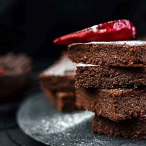 A stack of chocolate brownies dusted with powdered sugar is topped with a red pepper and placed on a dark plate.