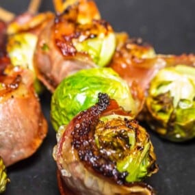 Close-up of skewered Brussels sprouts wrapped in crispy bacon, charred and roasted to a golden brown on a dark background.