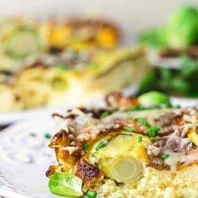 A close-up of a slice of vegetable frittata topped with cheese and garnished with chopped chives, served on a white plate. Other slices of frittata and vegetables are visible in the background.