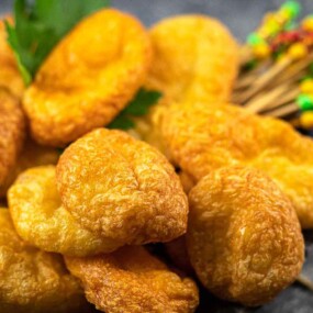A close-up of several golden-brown fried food items piled together with a skewer of colorful toothpicks and a garnish of green parsley.