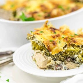 A slice of baked casserole containing broccoli, cheese, and other ingredients on a white plate, with the rest of the casserole in a white dish in the background. Two forks lie beside the plate.