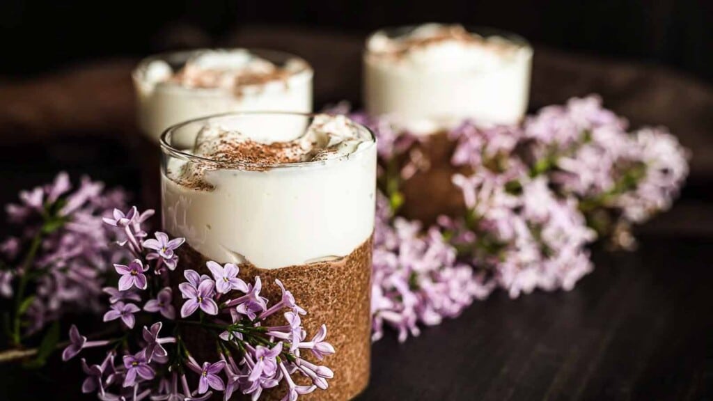 Three glass cups filled with a layered dessert topped with whipped cream and cocoa powder, surrounded by clusters of light purple flowers.