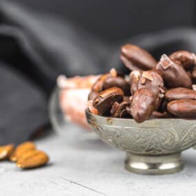 A silver cup filled with chocolate-covered almonds, placed on a light gray surface with more almonds and dark fabric in the background.