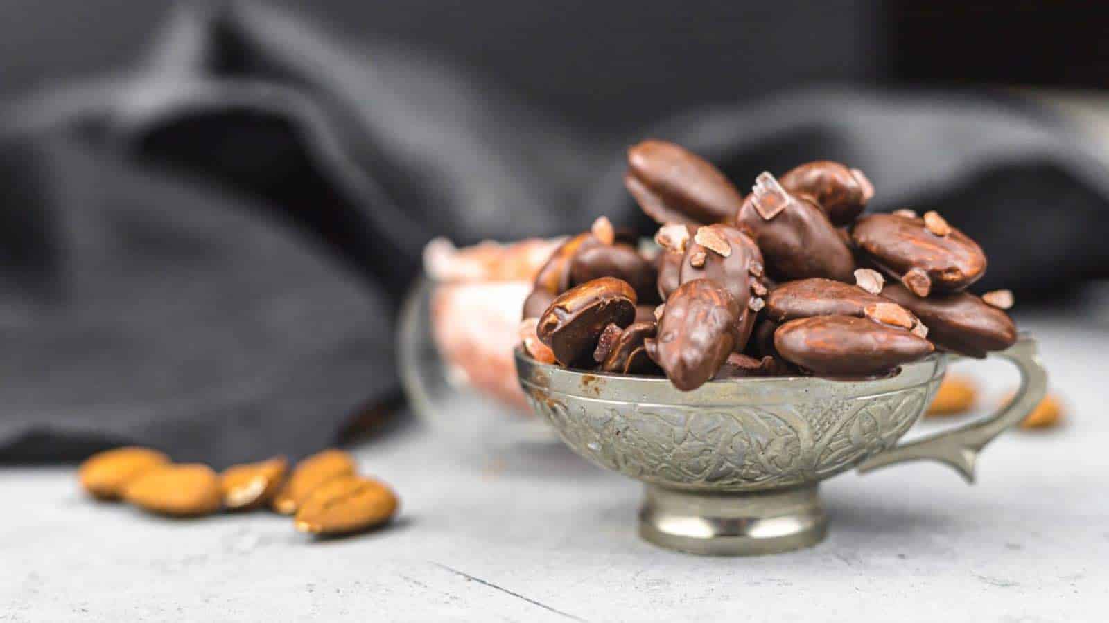 A silver cup filled with chocolate-covered almonds, placed on a light gray surface with more almonds and dark fabric in the background.