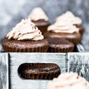 A group of chocolate cupcakes with brown frosting, arranged in a rustic wooden holder, against a blurred gray background.