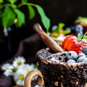 Close-up of a mug filled with a chocolate dessert topped with strawberries, blueberries, and a cinnamon stick, with another similar mug partially visible in the background, surrounded by plants and flowers.