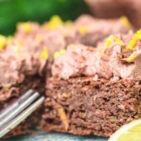 Close-up of a chocolate zucchini cake slice topped with chocolate frosting and lemon zest, with a fork and a slice of lemon nearby.