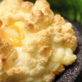 A golden-brown cloud egg with a soft yolk center is served on a dark stone plate, garnished with a green leaf.