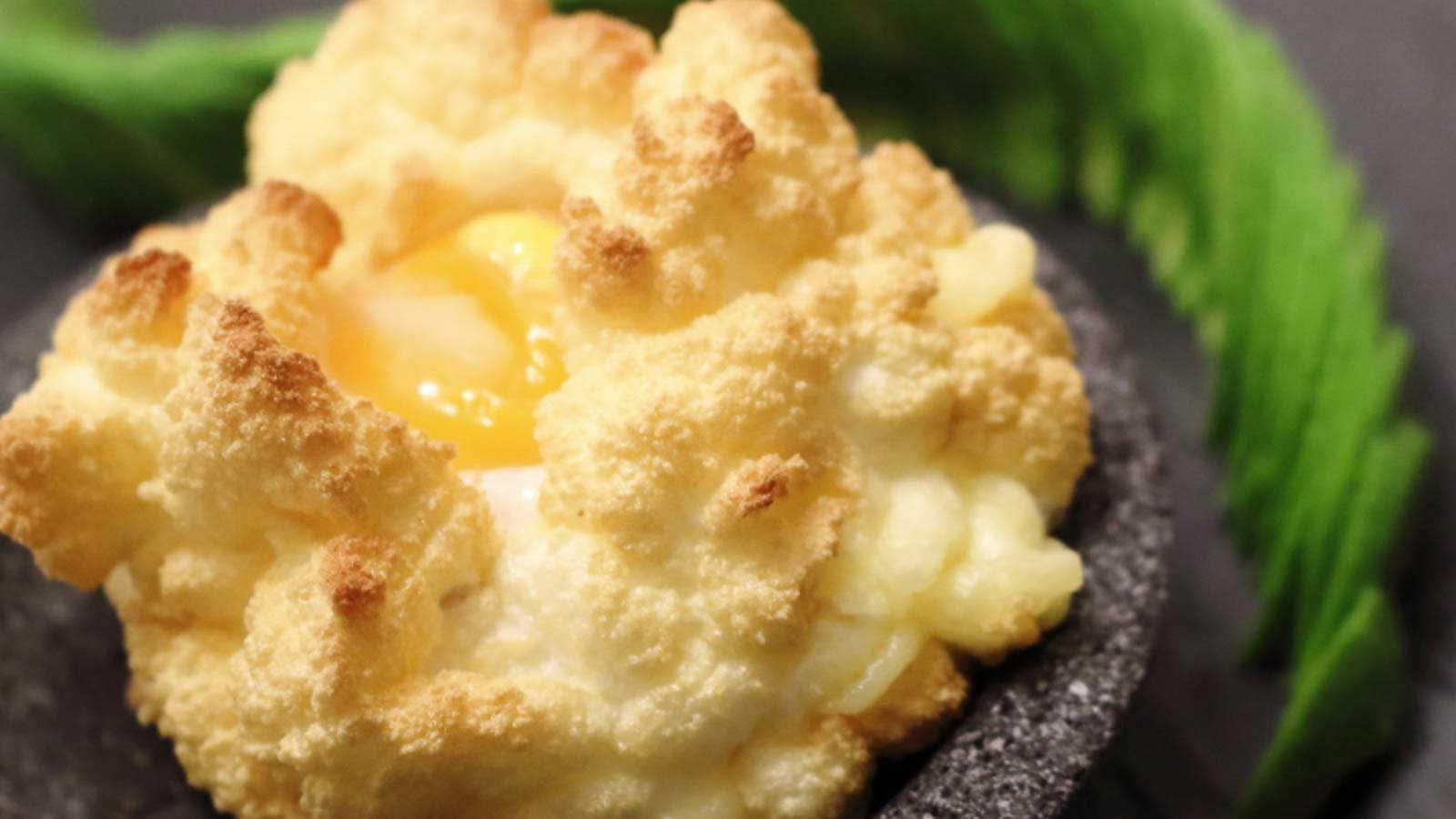 A golden-brown cloud egg with a soft yolk center is served on a dark stone plate, garnished with a green leaf.