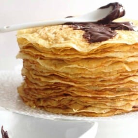 A stack of thin pancakes with a dollop of chocolate spread on top, placed on a white doily on a white surface.