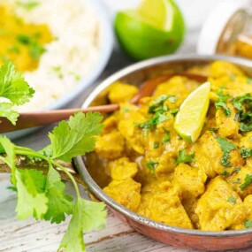 A bowl of chicken curry garnished with a lime wedge and cilantro, accompanied by a plate of rice, a lime half, and a jar of spices. Fresh cilantro is placed in the foreground.