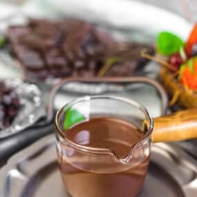 A small pot of melted chocolate on a plate, with a background featuring a dish of chocolate pieces, a bowl of berries, and a bowl of raisins.