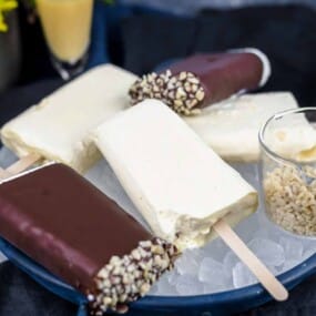 A plate of assorted ice cream bars, some covered in chocolate and nuts, others plain. A small glass cup filled with chopped nuts is also on the plate.