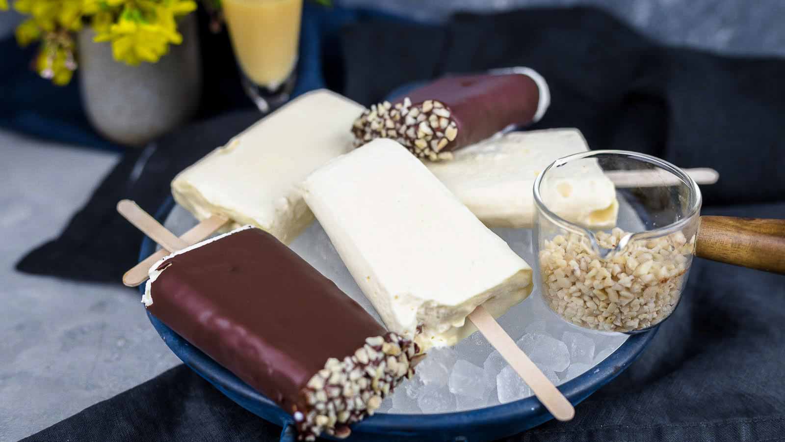 A plate of assorted ice cream bars, some covered in chocolate and nuts, others plain. A small glass cup filled with chopped nuts is also on the plate.
