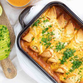 A dish of baked enchiladas covered in melted cheese and garnished with cilantro, accompanied by sliced avocado, lime wedges, and a small bowl of sauce.