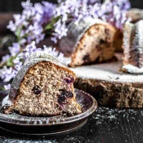 A slice of blueberry bundt cake on a plate, sprinkled with powdered sugar and decorated with purple flowers, with the remaining cake in the background on a wooden surface.
