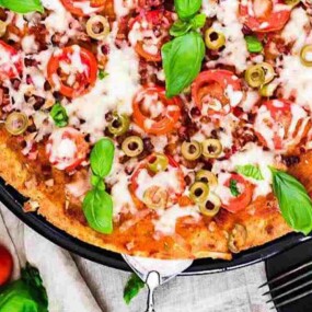 A freshly baked pizza topped with melted cheese, tomato slices, olives, basil leaves, and ground meat sits on a black tray. Nearby are cherry tomatoes, a small bowl of shredded cheese, and a fork.