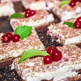 Close-up of dessert squares topped with white cream, red berries, and green mint leaves on a dark surface.