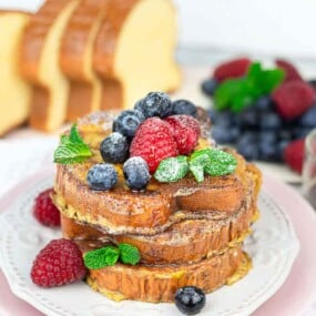 A dish of French toast topped with powdered sugar, mint leaves, raspberries, blueberries, and blackberries; maple syrup and sliced bread are in the background.