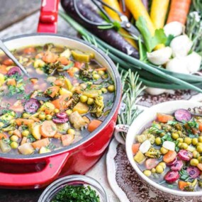 A colorful vegetable stew is served in a red pot with a silver spoon, accompanied by a bowl of stew and a variety of fresh vegetables and herbs on a rustic wooden table.
