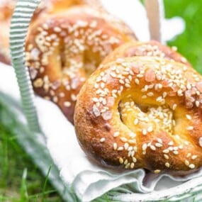 A wicker basket holds sesame seed-topped pretzels on a white cloth, placed on grass.