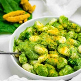 A white bowl filled with gnocchi covered in a green pesto sauce, garnished with yellow seasoning. A fork rests on the bowl's edge, and greens are in the background.