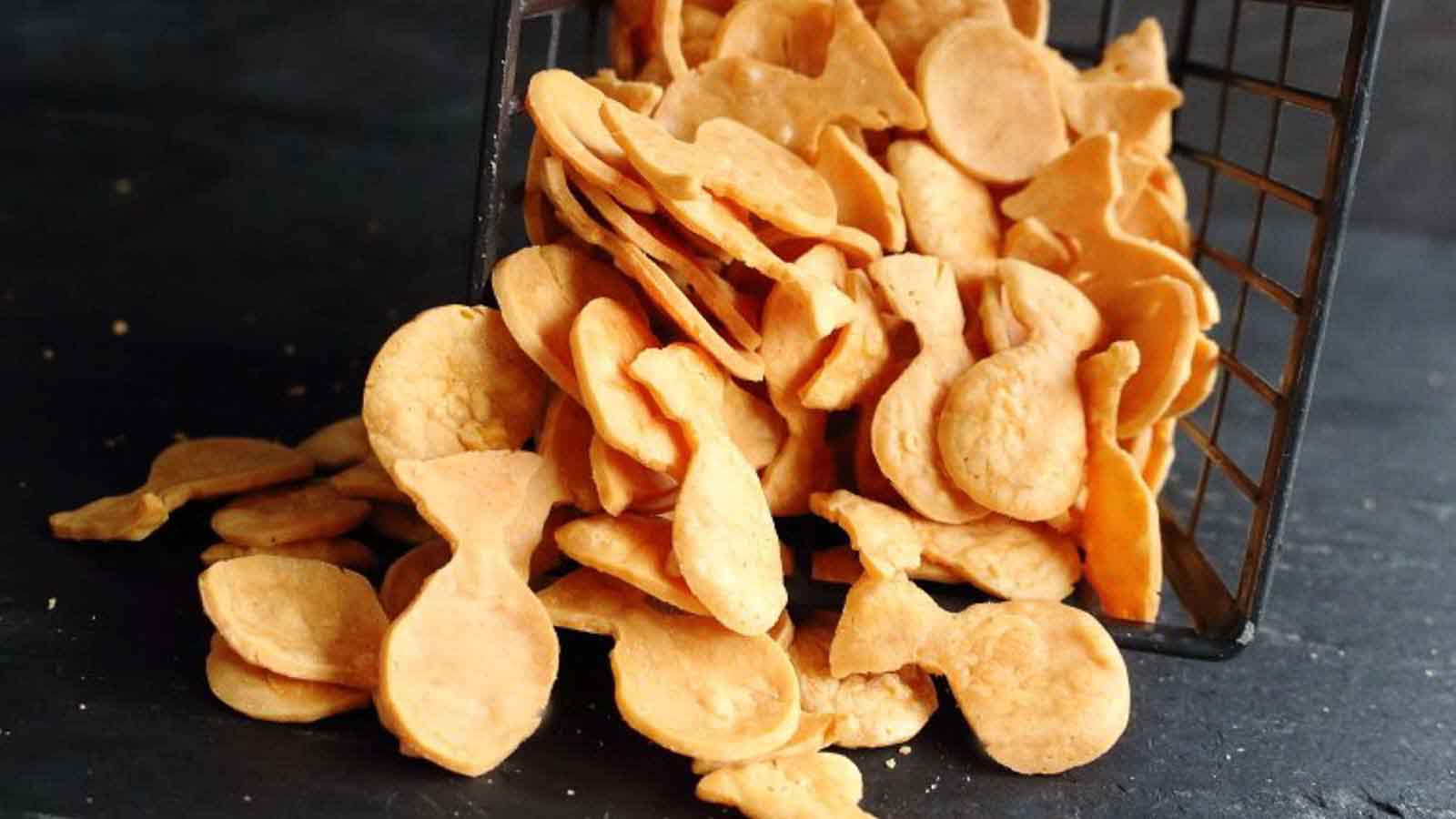 An overturned metal basket spilling out golden, fish-shaped crackers onto a dark surface.