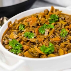 A white bowl filled with beef stew garnished with chopped parsley. The stew contains chunks of beef, sliced mushrooms, and diced carrots. A spoon is placed next to the bowl.