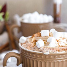 A close-up of a mug filled with hot chocolate topped with whipped cream and marshmallows, with another mug and a green salad bowl blurred in the background.