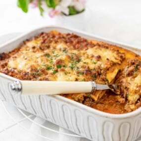 A baked lasagna in a rectangular white dish, with a portion missing and a serving utensil placed inside. The top layer is browned with melted cheese. A plant with small flowers is in the background.