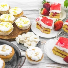A table features various desserts including cupcakes with white frosting and lemon zest, strawberry-topped cake squares, small meringues, fresh strawberries, and a potted plant.