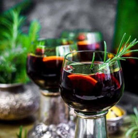 Three glasses of red sangria garnished with rosemary and fruit slices are on a table with scattered herbs and a green bottle in the background.