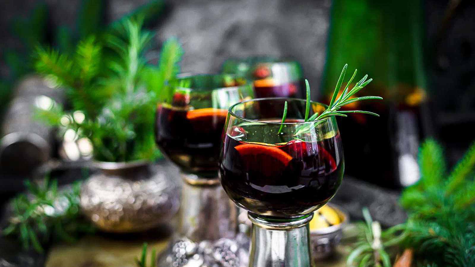 Three glasses of red sangria garnished with rosemary and fruit slices are on a table with scattered herbs and a green bottle in the background.