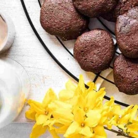 A black wire rack holds several chocolate cookies next to two bottles of milk and bright yellow flowers on a light wood surface.