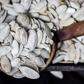 A close-up of a scoop filled with pumpkin seeds, with additional seeds filling a wooden container in the background.