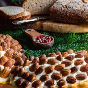 A cheese spread covered with almonds, arranged on a wooden board with a loaf of bread, sliced bread, a wooden spoon with mixed peppercorns, and pine branches for decoration.