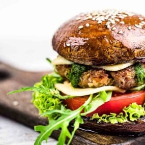 A gourmet burger with a mushroom cap bun, layered with a beef patty, cheese, pesto, lettuce, tomato, and arugula, served on a wooden board.