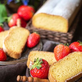 Slices of pound cake are arranged on plates beside fresh strawberries, with a loaf and additional strawberries in the background.