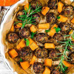 A rustic dish featuring meatballs, cubed butternut squash, and garnished with fresh rosemary and parsley. The meal is served on a parchment-lined plate on a wooden surface.