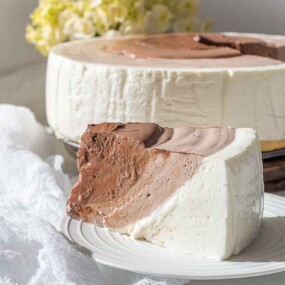 A slice of a layered chocolate and vanilla cheesecake is served on a white plate, with the rest of the cheesecake and a jar of cream in the background.