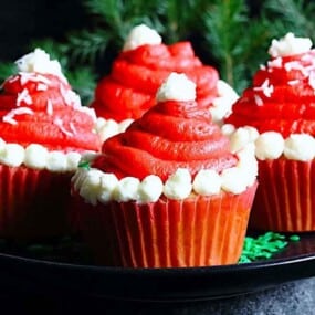 Four cupcakes with red frosting and white decoration resembling Santa hats are arranged on a black plate with greenery in the background.