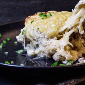 A serving of cheesy lasagna being lifted from a dark plate, garnished with chopped green herbs.